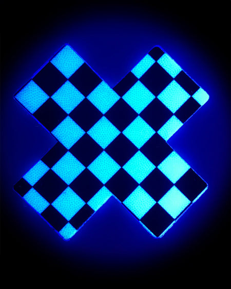 The Plus X: Black & White Checker Cross Nipple Pasties by Pastease shown glowing in the dark with a blue hue on a black background.