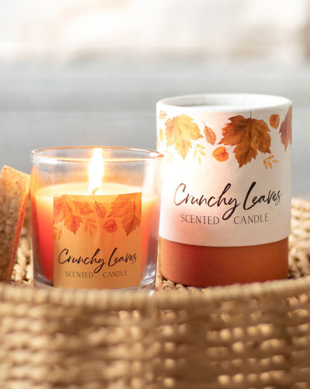 A close-up of the Rebel Romance Crunchy Leaves scented candle, flickering warmly in a glass jar with a cork lid. The packaging features delicate autumn leaves in earthy tones, creating a nostalgic seasonal atmosphere. Ideal for bringing cosy autumn scents indoors.