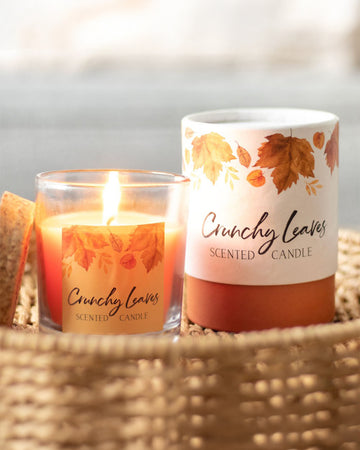 A close-up of the Rebel Romance Crunchy Leaves scented candle, flickering warmly in a glass jar with a cork lid. The packaging features delicate autumn leaves in earthy tones, creating a nostalgic seasonal atmosphere. Ideal for bringing cosy autumn scents indoors.