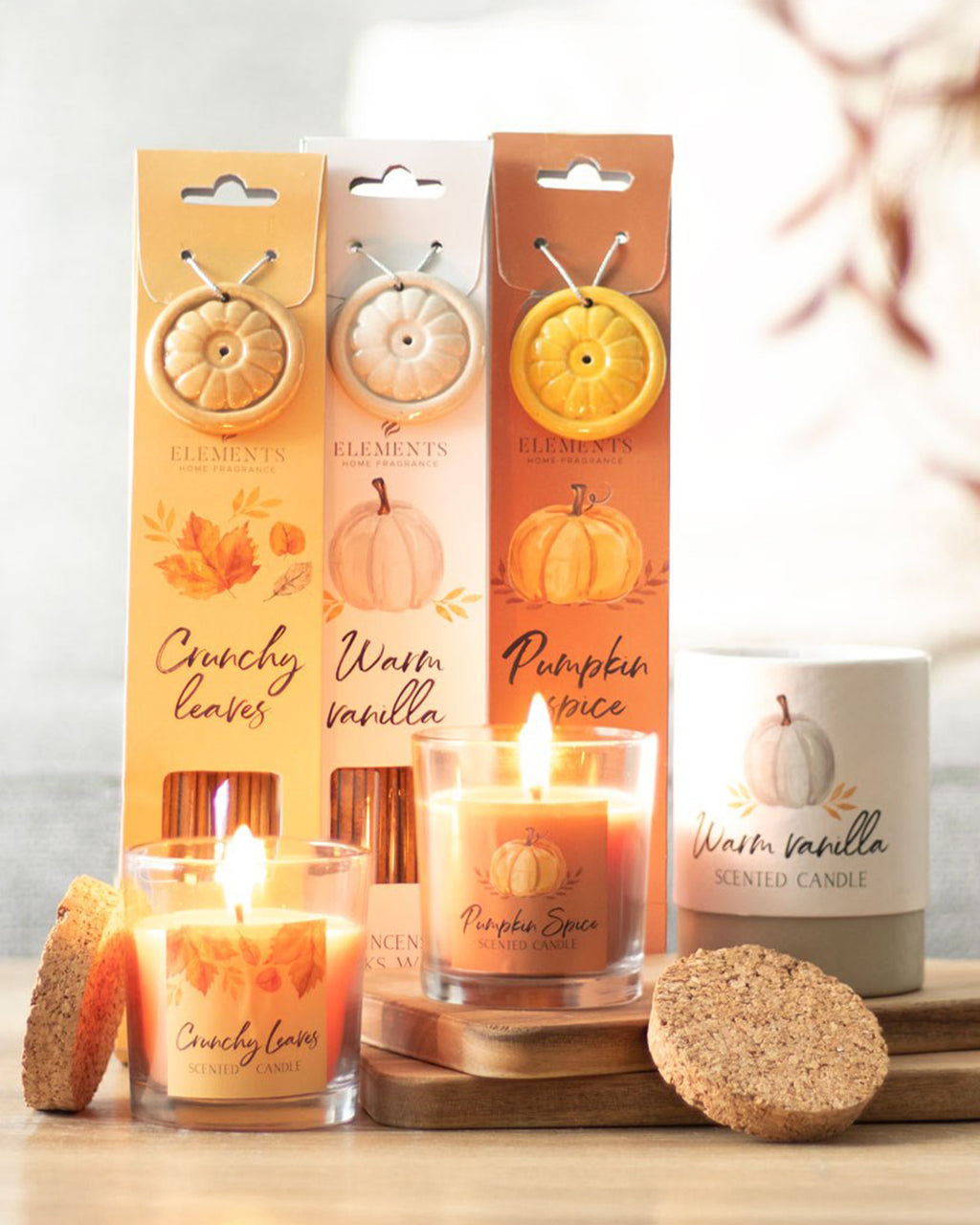 A cosy autumnal display of Rebel Romance's Crunchy Leaves, Warm Vanilla, and Pumpkin Spice candles, with matching scented ceramic ornaments in elegant packaging. Two lit candles sit on a wooden tray, casting a warm glow. A cork lid rests nearby, enhancing the rustic aesthetic. Perfect for embracing autumn vibes.