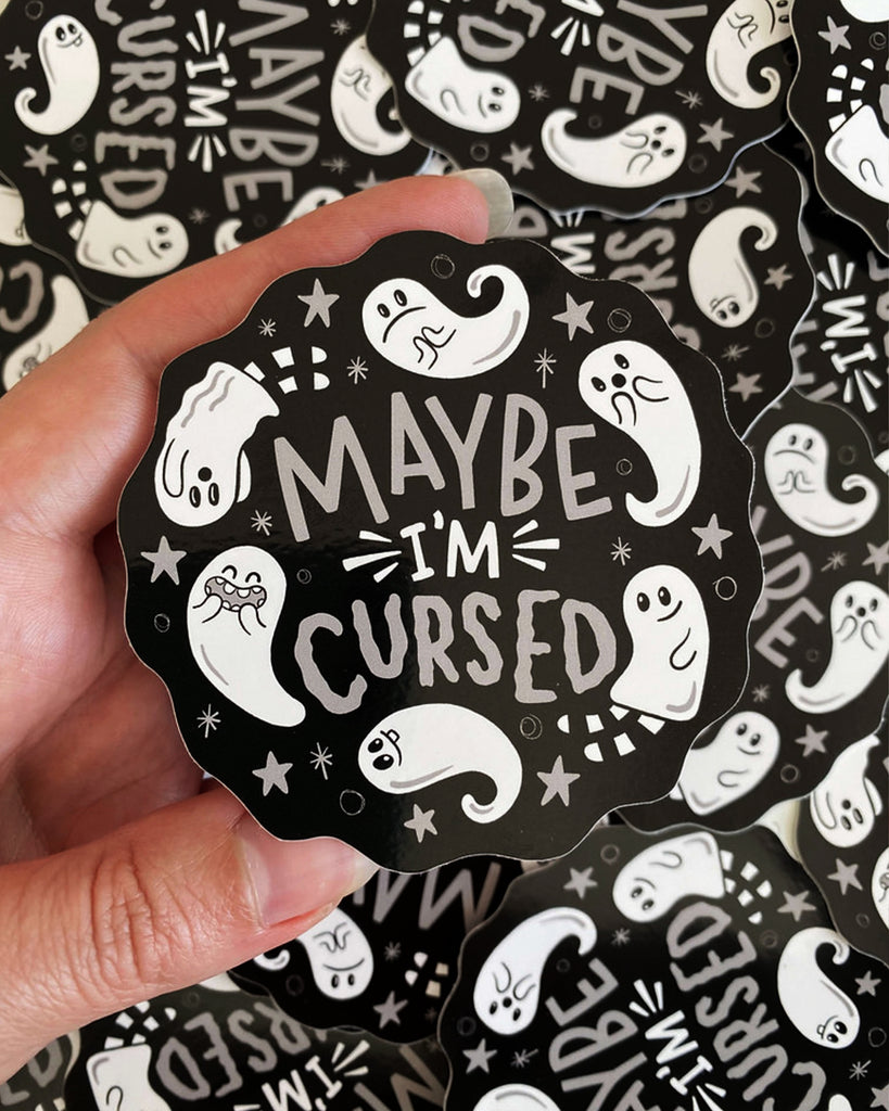 A hand holds a "Maybe I'm Cursed" ghost sticker from Rebel Romance, showing its full round shape with scalloped edges. The black sticker features white lettering and adorable ghosts illustrated by Yasmin Cousins, ideal for spooky, alternative style lovers.