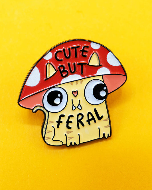 A close-up of the Cute But Feral mushroom cat enamel pin on a yellow background. The pin shows a wide-eyed yellow cartoon cat wearing a red toadstool mushroom hat, with the phrase “Cute But Feral” in bold black text. Designed by Katie Abey and stocked by Rebel Romance, a UK-based indie brand for lovers of playful, chaotic, and alternative accessories.