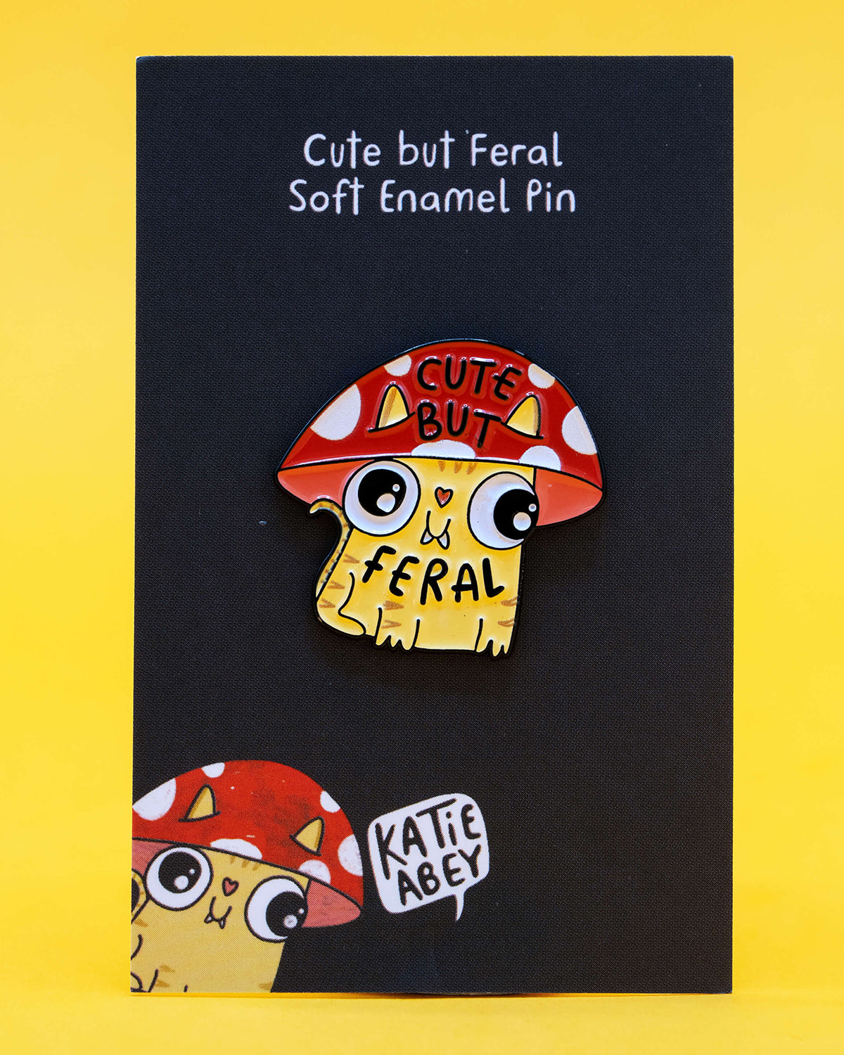 A soft enamel pin on a black backing card with white text that reads “Cute but Feral Soft Enamel Pin.” The pin features a yellow cartoon cat with a red and white toadstool mushroom hat, big eyes, and the words “Cute But Feral.” Illustrated by Katie Abey and sold by Rebel Romance, a UK alternative fashion and gift shop celebrating quirky, weird, and whimsical style.