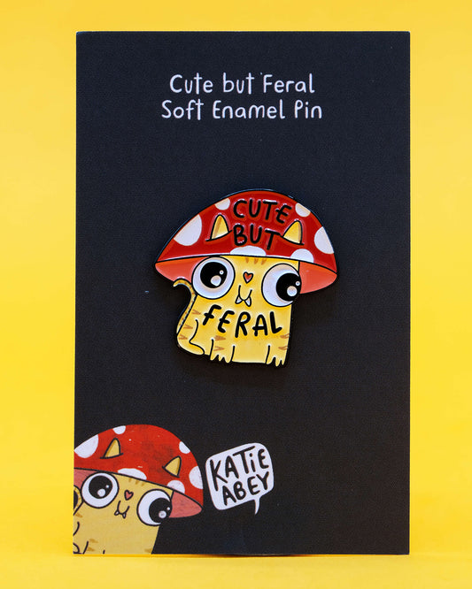 A soft enamel pin on a black backing card with white text that reads “Cute but Feral Soft Enamel Pin.” The pin features a yellow cartoon cat with a red and white toadstool mushroom hat, big eyes, and the words “Cute But Feral.” Illustrated by Katie Abey and sold by Rebel Romance, a UK alternative fashion and gift shop celebrating quirky, weird, and whimsical style.
