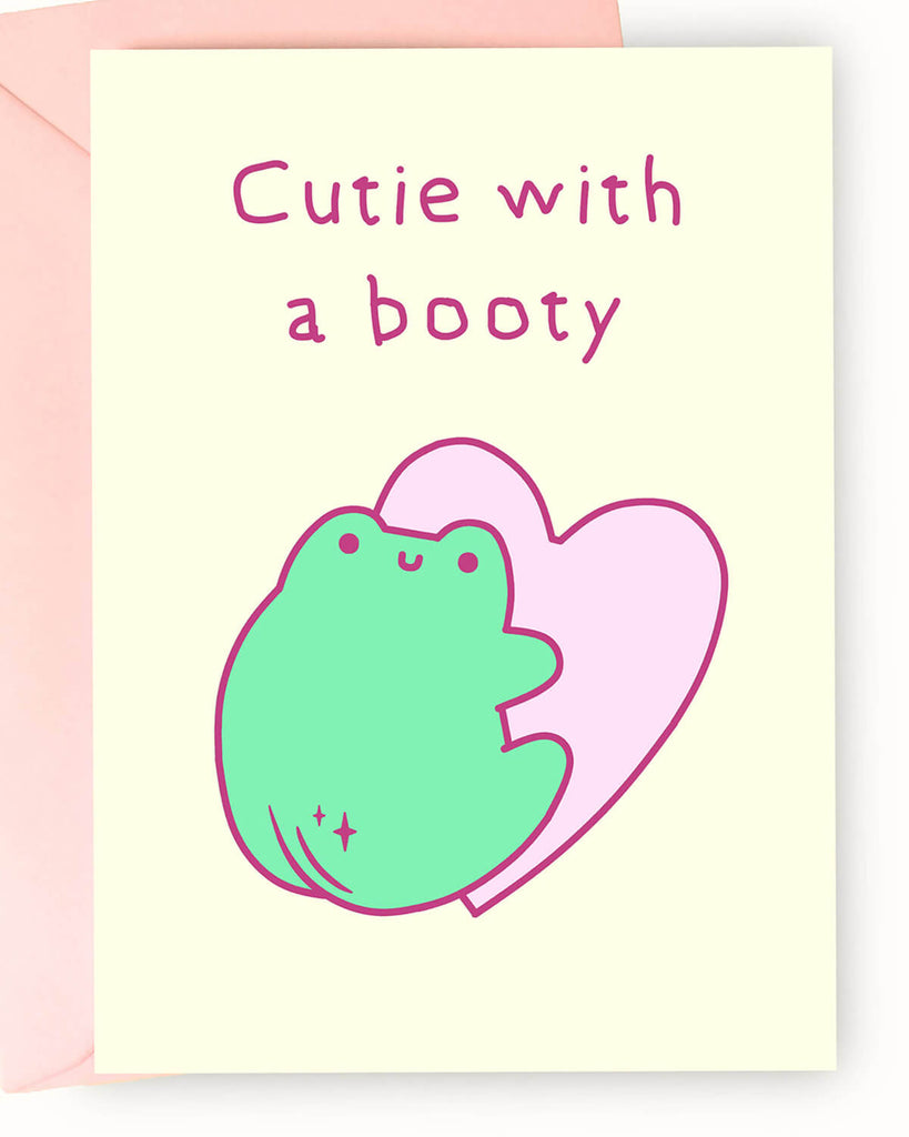 Adorable pastel greeting card from Rebel Romance featuring a cute green cartoon frog with a peachy booty in front of a pink heart, with the cheeky text “Cutie with a booty.” Set on a soft cream background and paired with a pale pink envelope, this playful card is perfect for quirky gifts, flirty notes, or spreading a little frog-loving sass. Ideal for millennials who love cute animals and fun alternative stationery.