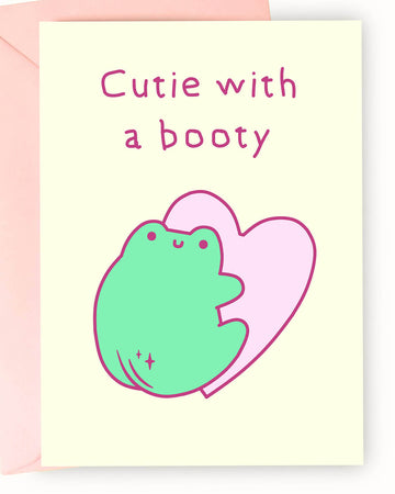 Adorable pastel greeting card from Rebel Romance featuring a cute green cartoon frog with a peachy booty in front of a pink heart, with the cheeky text “Cutie with a booty.” Set on a soft cream background and paired with a pale pink envelope, this playful card is perfect for quirky gifts, flirty notes, or spreading a little frog-loving sass. Ideal for millennials who love cute animals and fun alternative stationery.