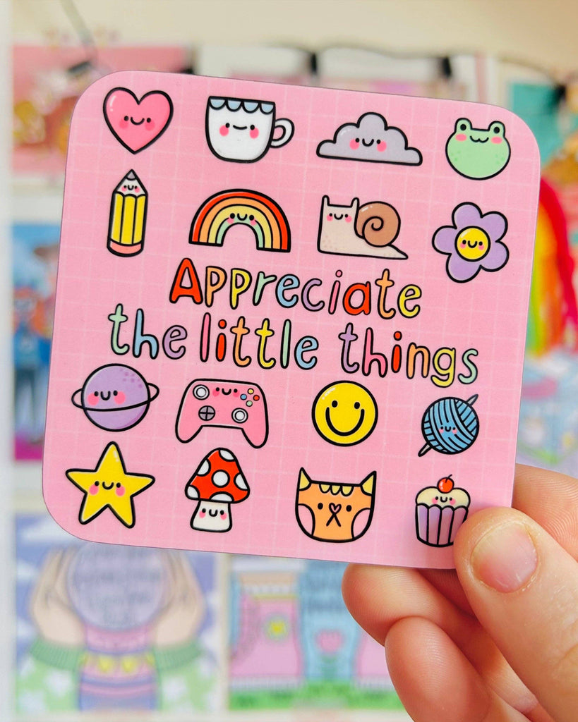 The Appreciate The Little Things drinks coaster by Kelly Lou from Rebel Romance, held in hand. The pink coaster is covered in fun kawaii‑style illustrations such as a star, pencil, cupcake and rainbow, alongside bold multicoloured text. A perfect alternative gift celebrating small joys and cute homeware.