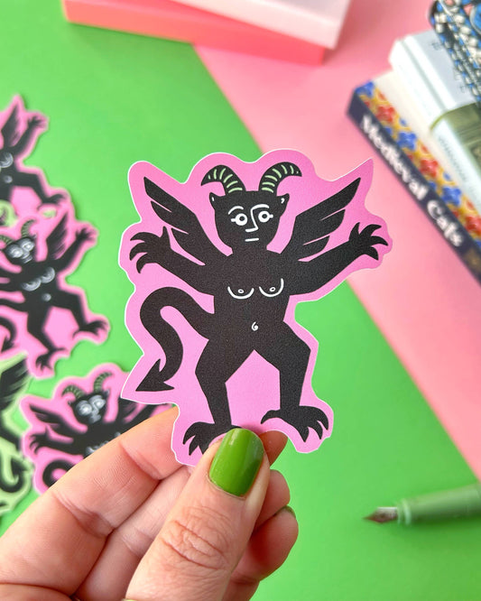 Close-up of a person holding a Pink Devil Sticker by Stacey McEvoy Caunt in front of a bright green and pink background. The devil has six arms, wings, horns, and a cheeky expression, printed in black on a pink vinyl background. Other stickers and a fountain pen are visible in the background. Sold by Rebel Romance, a UK brand celebrating alternative style.