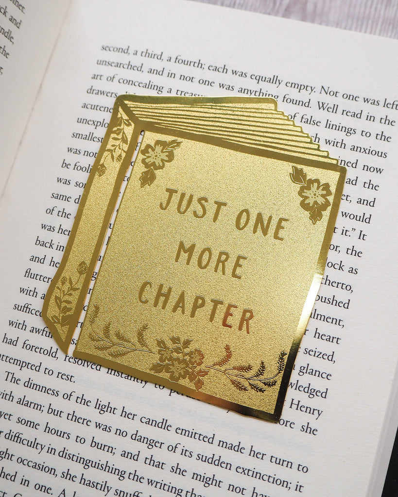 A detailed Literary Emporium brass bookmark engraved with flowers and the phrase “Just One More Chapter,” styled on an open page. Sold by Rebel Romance, a UK indie brand offering alternative gifts, homeware and slow ethical fashion.