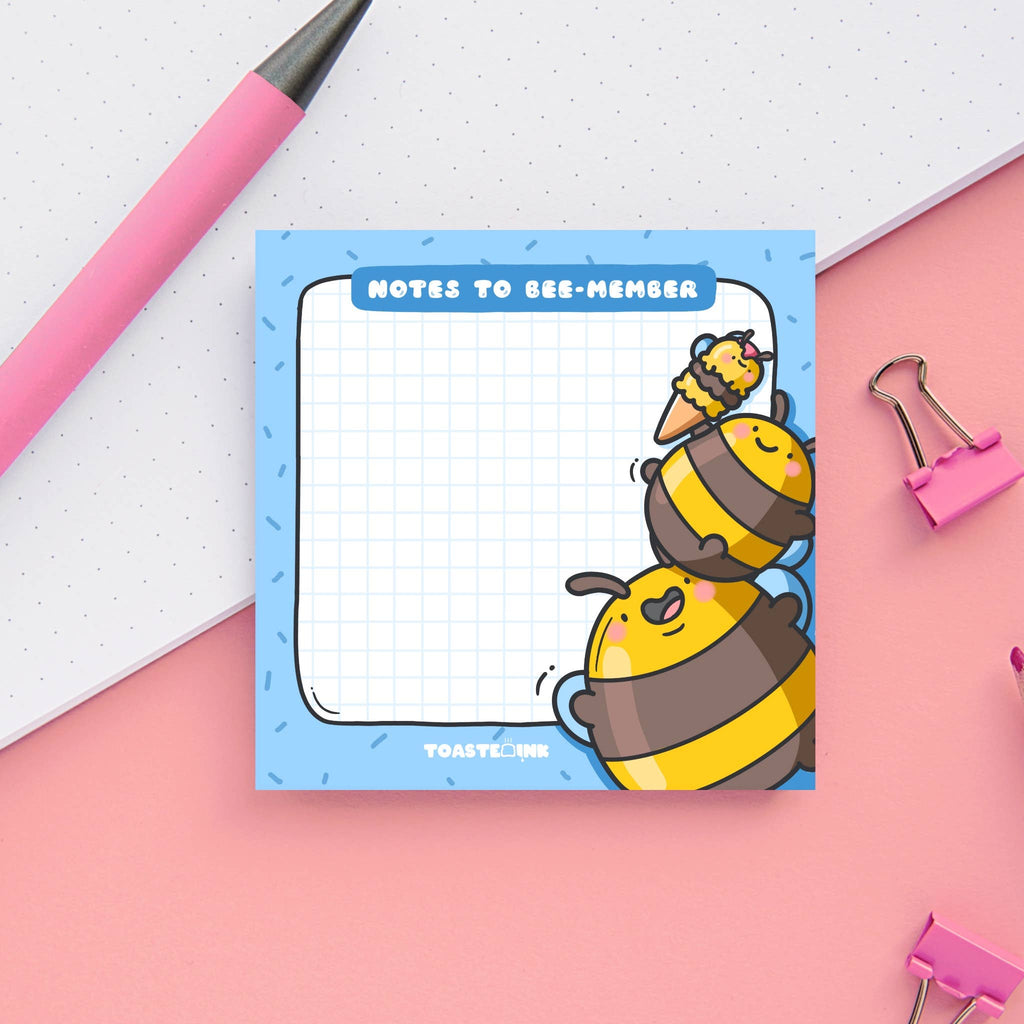 Square sticky note pad with a blue border and grid paper, featuring two chubby yellow and brown cartoon bumble bees stacking on top of each other to reach an ice cream. The top reads “Notes to Bee-member” in bold white text. Cute and practical stationery for daily reminders.