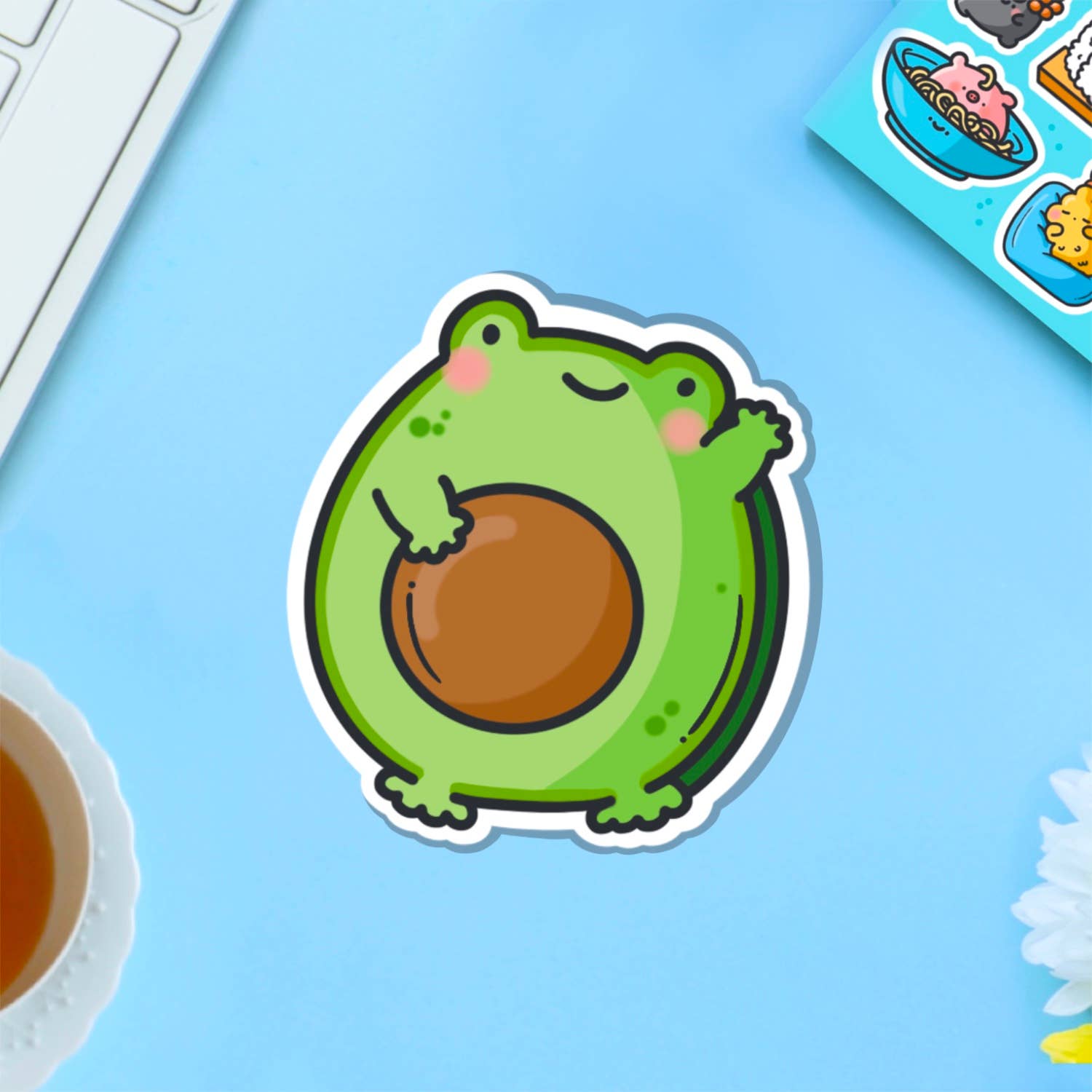 A cute vinyl sticker of a green cartoon frog with rosy cheeks, shaped like an avocado with a large brown stone in the centre. The sticker is shown on a purple background. Ideal for decorating laptops, water bottles, notebooks and more.