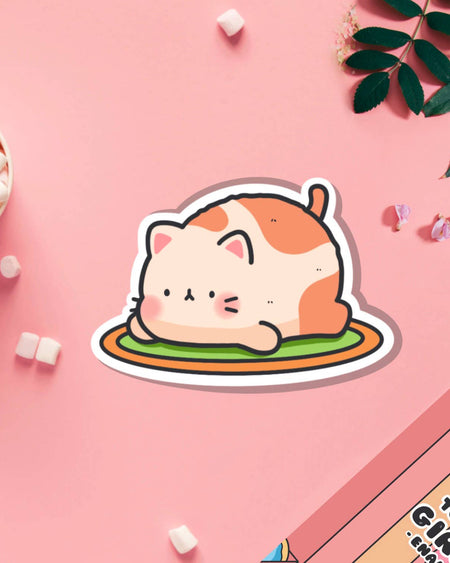 Adorable Resting Cat Vinyl Sticker by Toastedink, sold by Rebel Romance. A cute chubby cat with peach and orange markings lounges on a green rug, placed on a pastel pink surface with marshmallows and leaves. A perfect gift for cat lovers and alternative stationery fans in the UK.