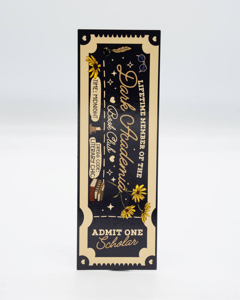 Black and gold ouija board-style book displayed with a dark academia-themed bookmark from Rebel Romance. The cream and black ticket-style bookmark reads “Lifetime Member of the Dark Academia Book Club – Admit One Scholar.” Designed by UK brand Rebel Romance, known for alternative, millennial-friendly gifts, homeware, and slow fashion with a gothic twist.
