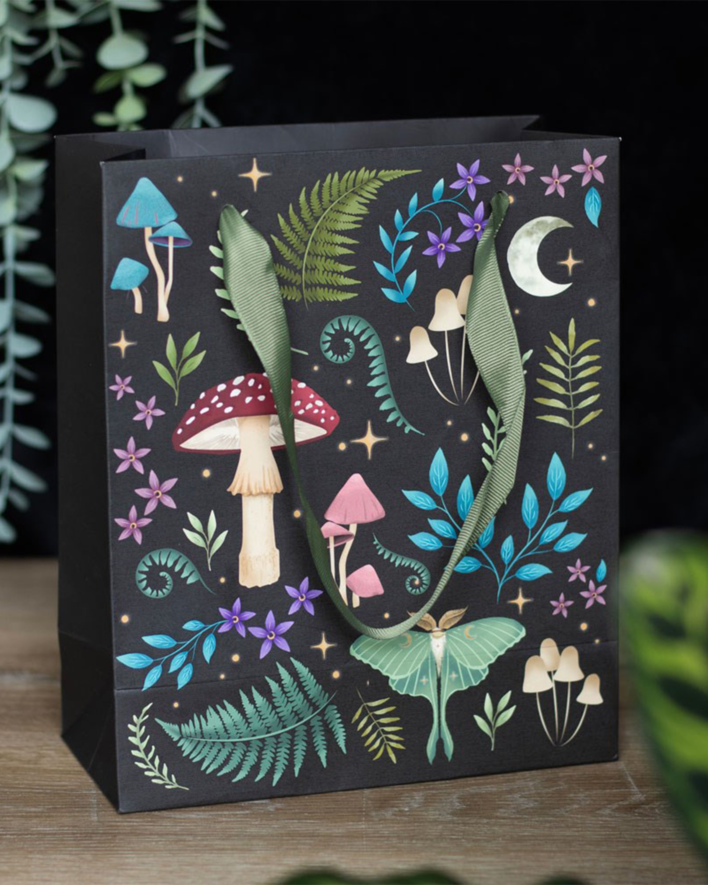 Rebel Romance Medium Dark Forest Print Gift Bag photographed on a wooden surface, with green ribbon handles and a dark botanical print of mushrooms, ferns, flowers and a luna moth, perfect for alternative style lovers and magical gifts.