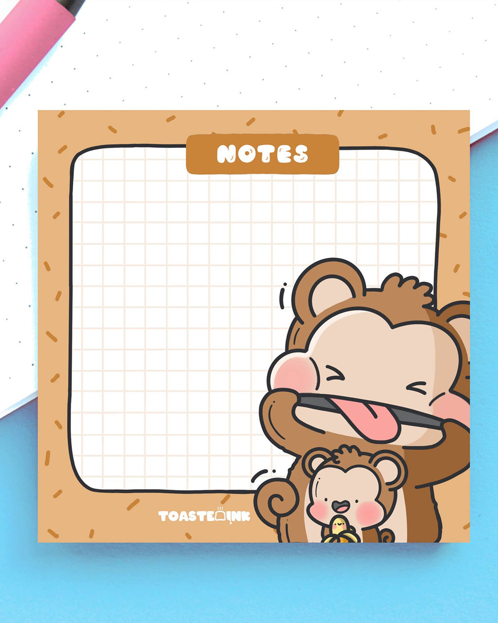 Flat lay of Toastedink monkey sticky notes on a blue background with a pink pen. Features cheeky cartoon monkeys and a gridded note space. Available at Rebel Romance, a UK indie store for whimsical and retro stationery lovers.