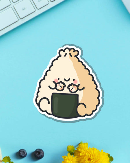 Adorable Toastedink vinyl sticker of a dumpling onigiri character holding mini dumplings, placed on a pastel blue background with yellow flowers and a keyboard. Stocked by Rebel Romance, a UK brand celebrating fun and alternative aesthetic gifts.