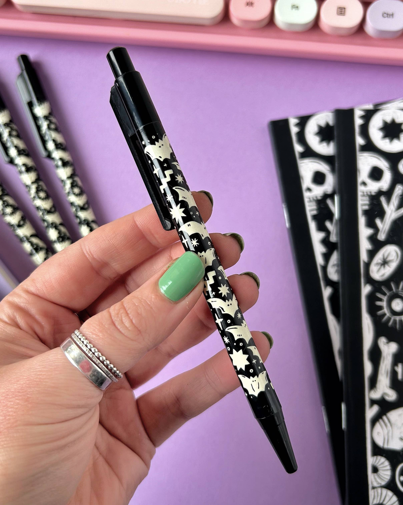 A Rebel Romance Bat Pattern Pen held in hand over a purple surface, surrounded by matching gothic notebooks and other pens, showcasing the full illustrated wraparound design.