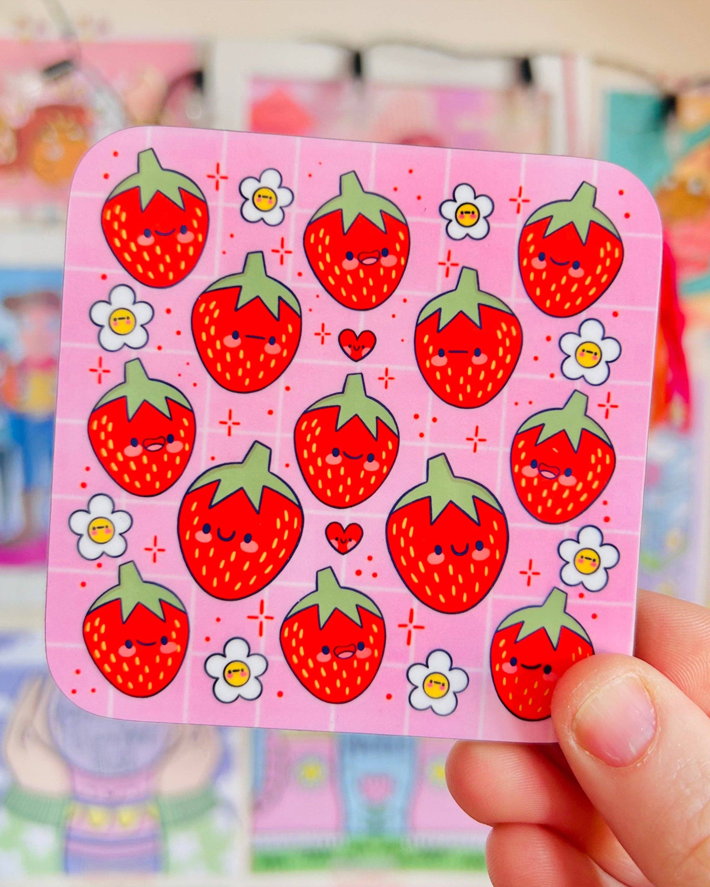 A hand holding the Strawberry Cuties Drinks Coaster by Kelly Lou from Rebel Romance. The square pink coaster features cute illustrated strawberries with smiling faces and tiny flowers, making it a fun, alternative home accessory or gift for lovers of unique design.