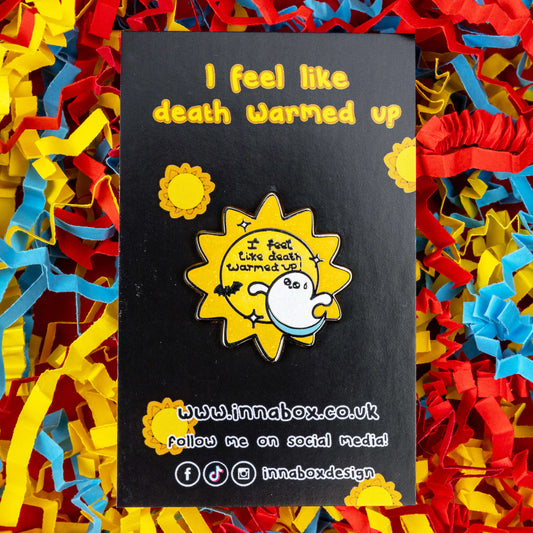 Death Warmed Up ghost enamel pin on a black Innabox backing card with yellow text and sun icons, styled on red, blue, and yellow shredded paper. The Halloween-themed pin reads “I feel like death warmed up!” and features a sparkly yellow sun, ghost, and black bat. A playful enamel pin from Rebel Romance for spooky vibes and chronic illness awareness.