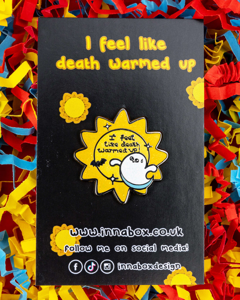 Death Warmed Up ghost enamel pin on a black Innabox backing card with yellow text and sun icons, styled on red, blue, and yellow shredded paper. The Halloween-themed pin reads “I feel like death warmed up!” and features a sparkly yellow sun, ghost, and black bat. A playful enamel pin from Rebel Romance for spooky vibes and chronic illness awareness.
