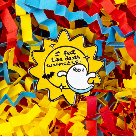 Close-up of Rebel Romance’s Death Warmed Up glitter enamel pin, designed by Innabox, surrounded by colourful shredded paper in red, blue, and yellow. The pin features a cute ghost and bat with the phrase “I feel like death warmed up!” on a yellow glittery sunburst background. Perfect for alternative fashion lovers and Halloween gifts.