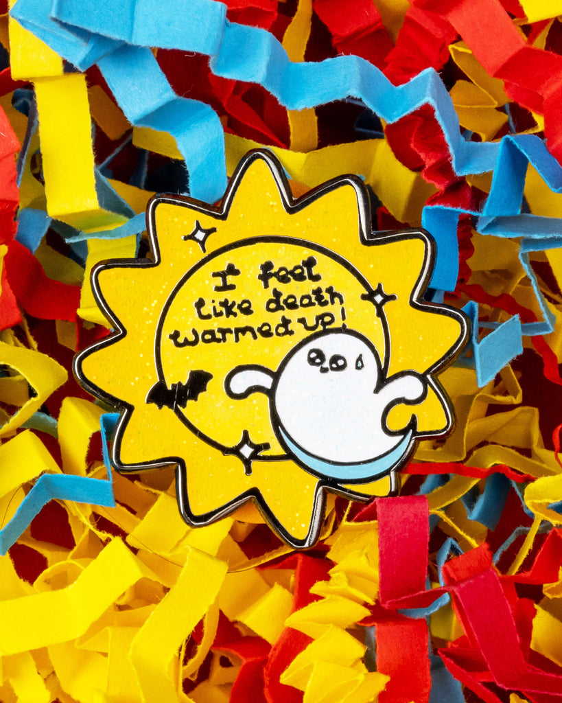 Close-up of Rebel Romance’s Death Warmed Up glitter enamel pin, designed by Innabox, surrounded by colourful shredded paper in red, blue, and yellow. The pin features a cute ghost and bat with the phrase “I feel like death warmed up!” on a yellow glittery sunburst background. Perfect for alternative fashion lovers and Halloween gifts.