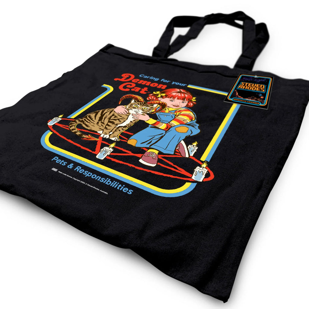 A black tote bag featuring a Steven Rhodes design titled “Demon Cat.” The artwork shows a young girl sitting with a horned tabby cat in a pentagram surrounded by lit candles. The text “Caring for your Demon Cat” and “Pets & Responsibilities” appear above and below the design. The colourful, retro-style illustration contrasts with the dark background.
