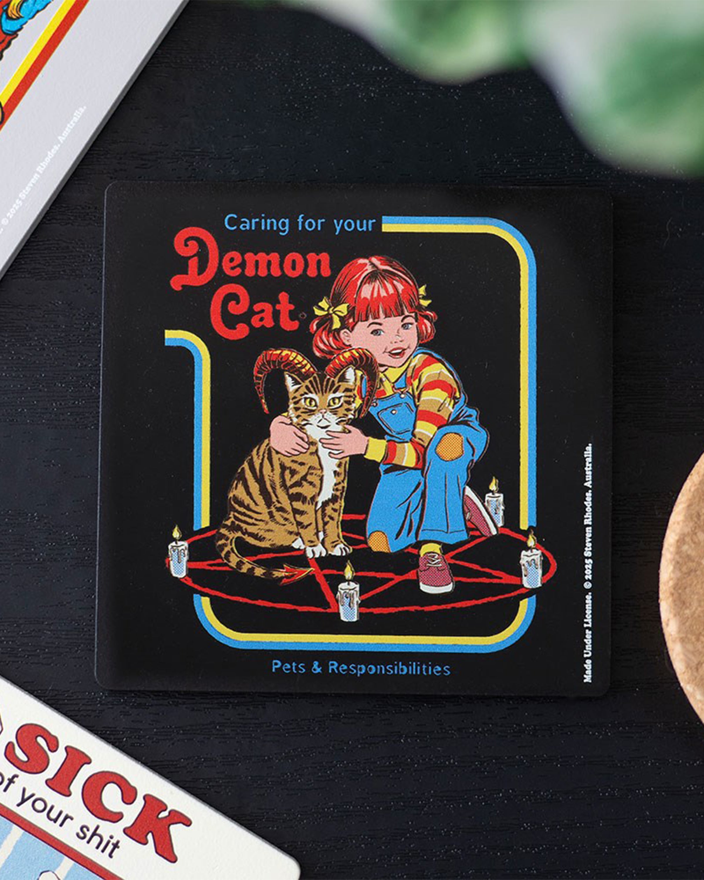 Styled shot of the Caring for Your Demon Cat Steven Rhodes coaster on a table, available at Rebel Romance. Features retro horror-inspired artwork with a girl and her demon cat inside a summoning circle. Ideal alternative homeware gift.
