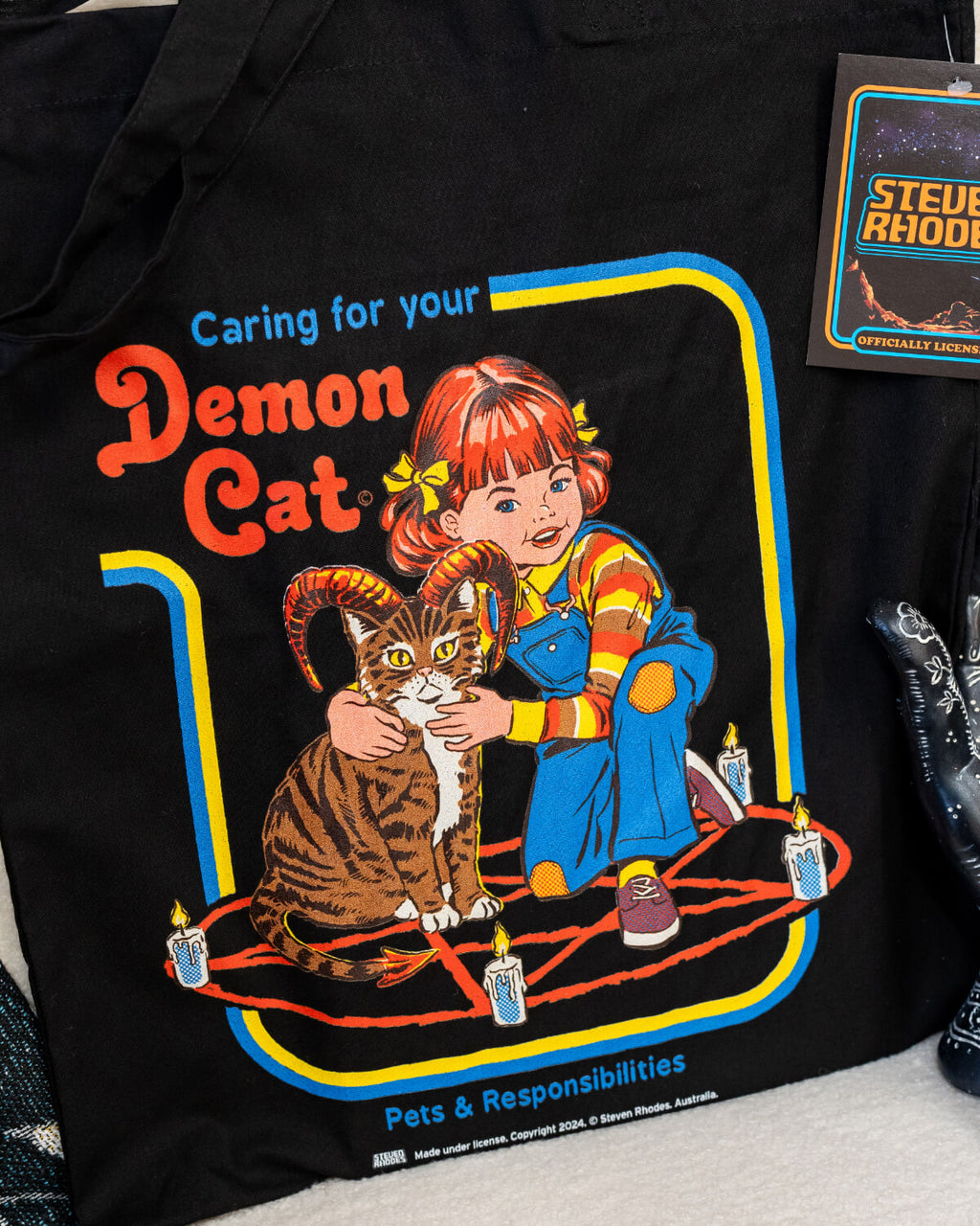 A black tote bag featuring a Steven Rhodes design titled “Demon Cat.” The artwork shows a young girl sitting with a horned tabby cat in a pentagram surrounded by lit candles. The text “Caring for your Demon Cat” and “Pets & Responsibilities” appear above and below the design. The colourful, retro-style illustration contrasts with the dark background.