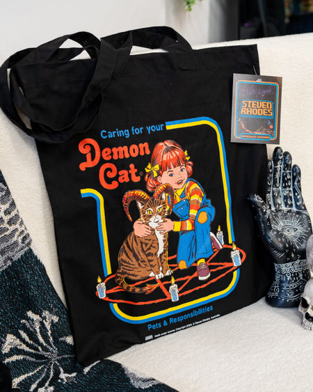 A black tote bag featuring a Steven Rhodes design titled “Demon Cat.” The artwork shows a young girl sitting with a horned tabby cat in a pentagram surrounded by lit candles. The text “Caring for your Demon Cat” and “Pets & Responsibilities” appear above and below the design. The colourful, retro-style illustration contrasts with the dark background.