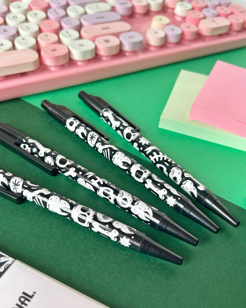 Four black ballpoint pens with a white skull and bones pattern, laid diagonally on a green background beside a pastel pink keyboard and colourful sticky notes. These gothic stationery pens are from Rebel Romance, a small UK brand celebrating alternative style.