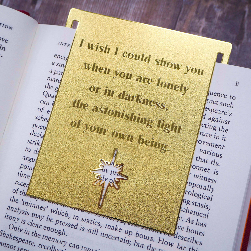A close-up image of a brass bookmark engraved with the heartfelt Hafiz of Shiraz quote: “I wish I could show you, when you are lonely or in darkness, the astonishing light of your own being.” The bookmark is resting on the pages of an open book, showcasing its intricate detailing and elegant design.