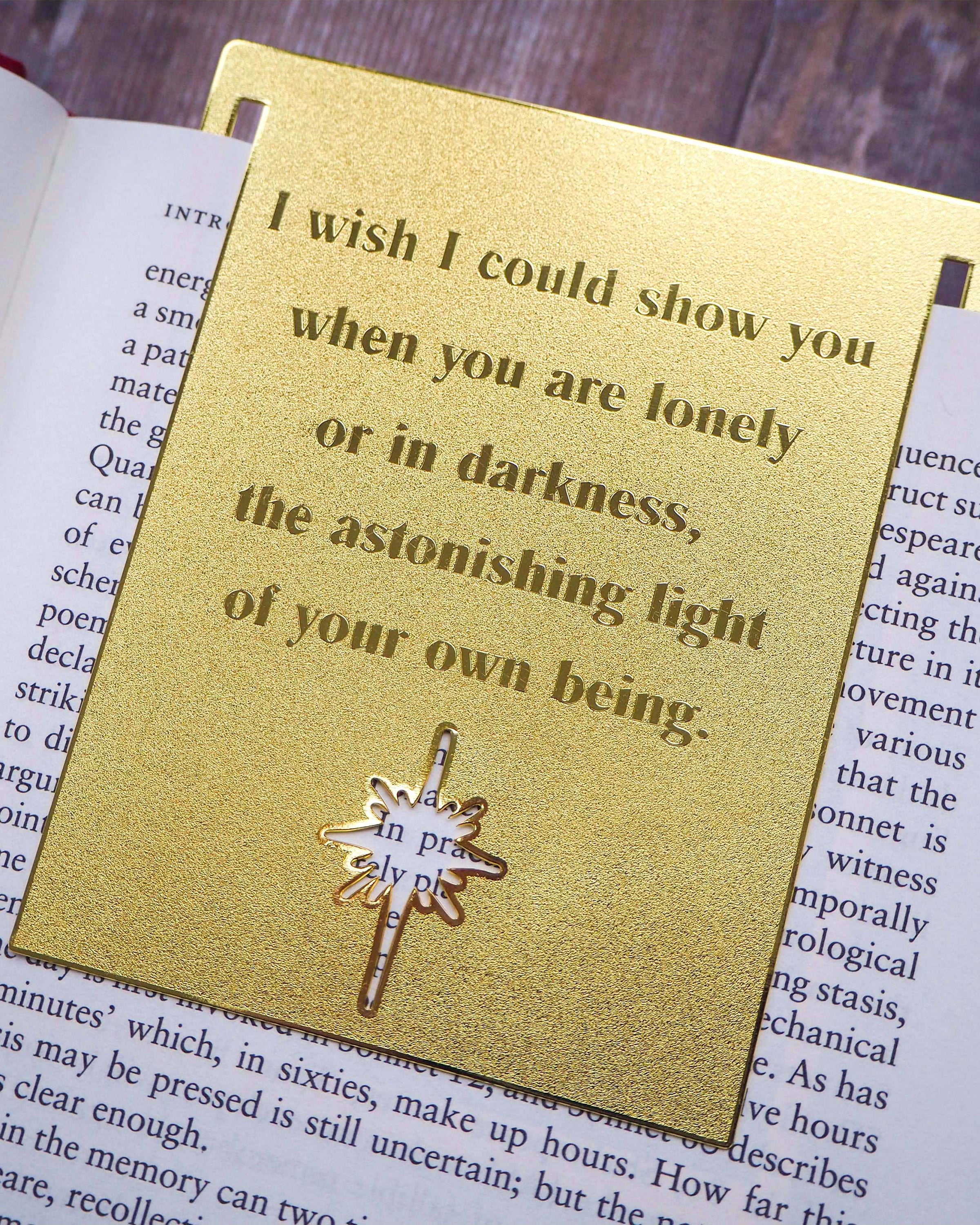 A close-up image of a brass bookmark engraved with the heartfelt Hafiz of Shiraz quote: “I wish I could show you, when you are lonely or in darkness, the astonishing light of your own being.” The bookmark is resting on the pages of an open book, showcasing its intricate detailing and elegant design.