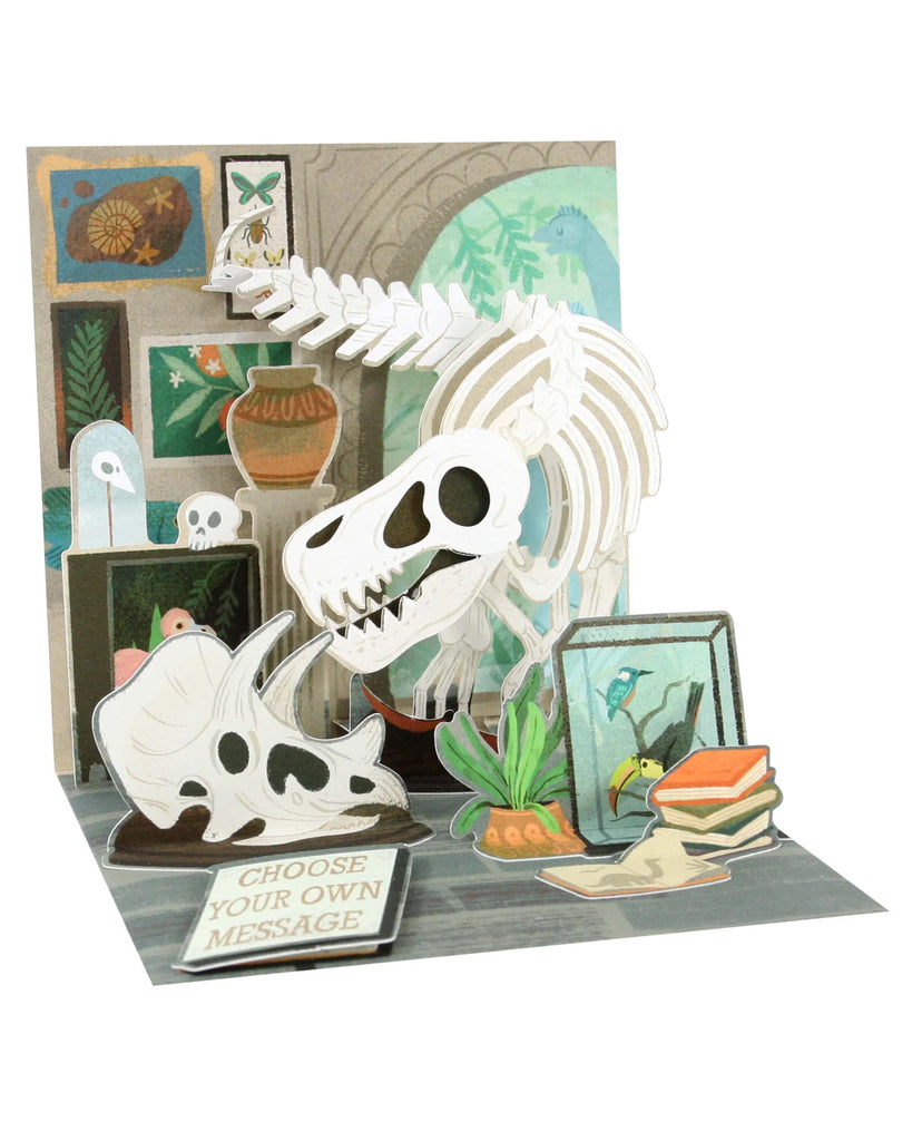 A colourful pop-up greeting card featuring a 3D dinosaur skeleton surrounded by fossils, plants, and books in a museum-style setting. The card includes interchangeable tags with messages such as “You Are Dinomite!” and “Happy Birthday.” Finished with copper foil and comes with a white envelope, perfect for science lovers and dinosaur enthusiasts.