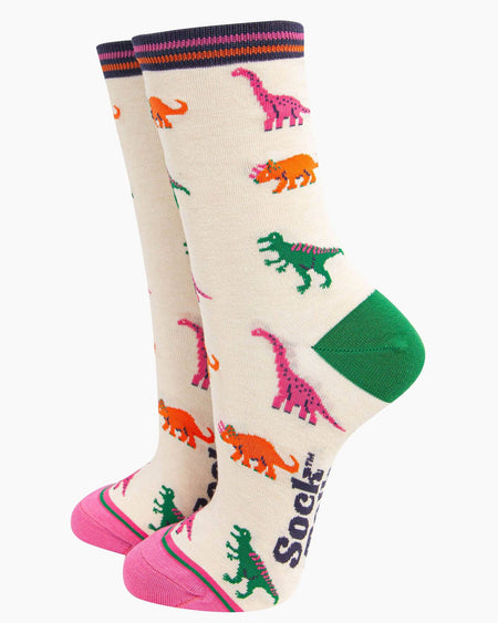 A pair of cream bamboo socks with a playful dinosaur print featuring green T-Rex, pink brachiosaurus, and orange triceratops, finished with contrasting pink toes, green heels, and a striped cuff.