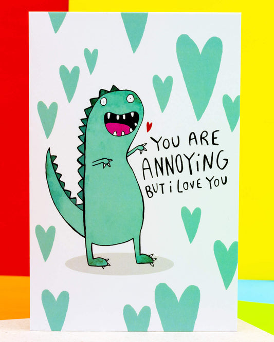 A colourful A6 greeting card by Rebel Romance featuring a cute green dinosaur illustration by Katie Abey with the caption "You Are Annoying But I Love You" in playful handwritten text, surrounded by mint green hearts. A cheeky and affectionate card perfect for Valentine's Day, anniversaries, or just because. Made in the UK.