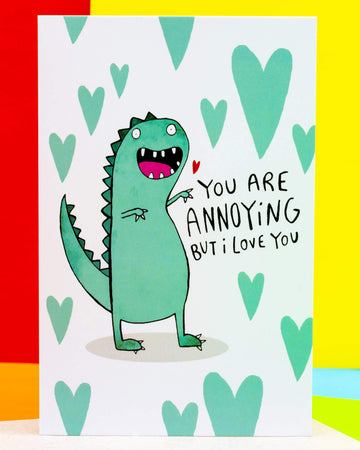A colourful A6 greeting card by Rebel Romance featuring a cute green dinosaur illustration by Katie Abey with the caption "You Are Annoying But I Love You" in playful handwritten text, surrounded by mint green hearts. A cheeky and affectionate card perfect for Valentine's Day, anniversaries, or just because. Made in the UK.