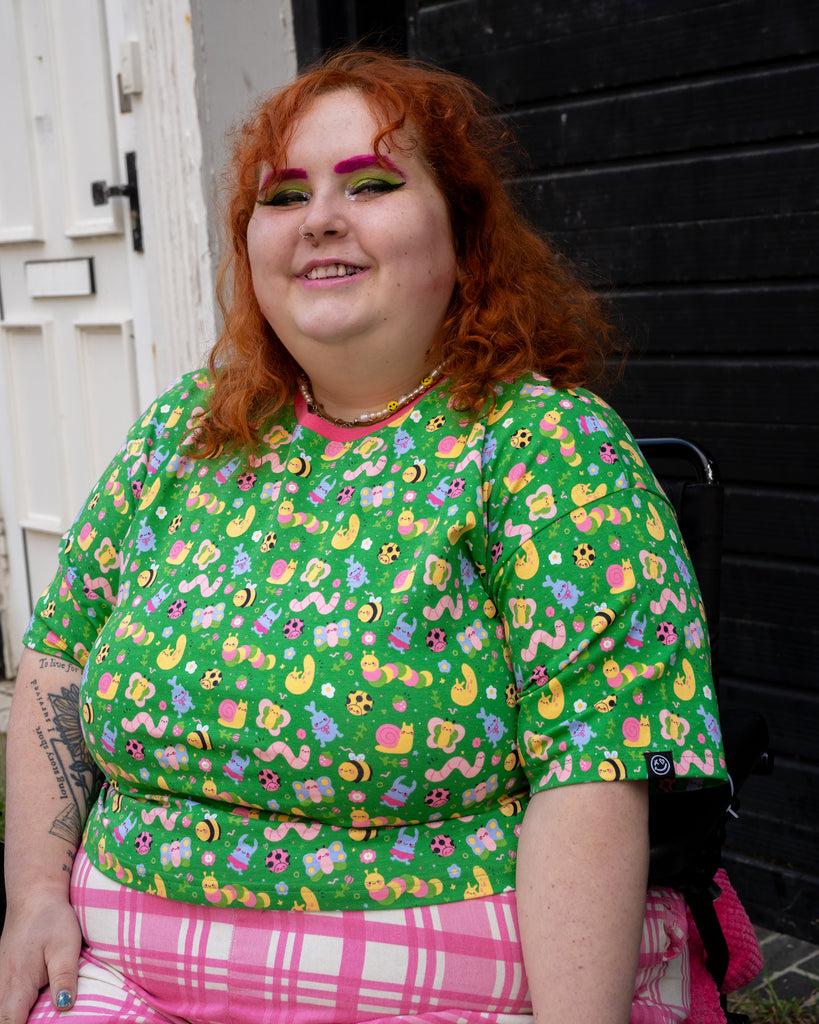 Lauren wears the Rebel Romance x Dipstick Store Bugs Cropped Boxy Tee and sits outside on a street with black and white buildings. The green top features a colourful bug print with a pink collar and soft stretchy fit. Ethical alternative fashion by Rebel Romance, made for dopamine dressers.
