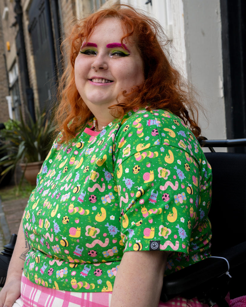 Side view of Lauren wearing the Rebel Romance x Dipstick Store Bugs Cropped Boxy Tee. The top features a cheerful print of snails, worms, moons, and ladybirds on a bright green background, paired with pink check trousers. Inclusive fashion by UK brand Rebel Romance.
