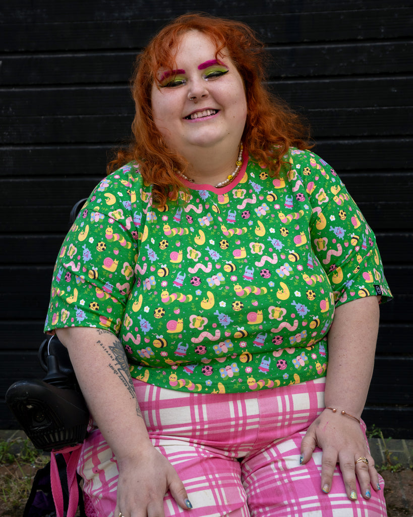 Lauren, a size 22/24 model in a wheelchair, smiles brightly while wearing the Rebel Romance x Dipstick Store Bugs Cropped Boxy Tee. The bold green tee features pastel bugs, flowers, and worms, styled with pink plaid trousers. A colourful look from UK slow fashion brand Rebel Romance.