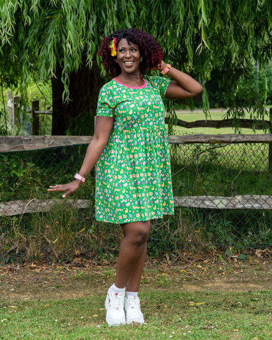 Ayesha (she/her), 5'10" and a UK size 8/10, poses in the Rebel Romance x Dipstick Store Bugs Stretch Smock Dress, wearing a size 2XS. This colourful green dress with an all-over bug print and pink neckline is part of Rebel Romance’s playful, ethical fashion range. Photographed in a grassy field with wooden fencing and trees, this look is perfect for dopamine dressers.