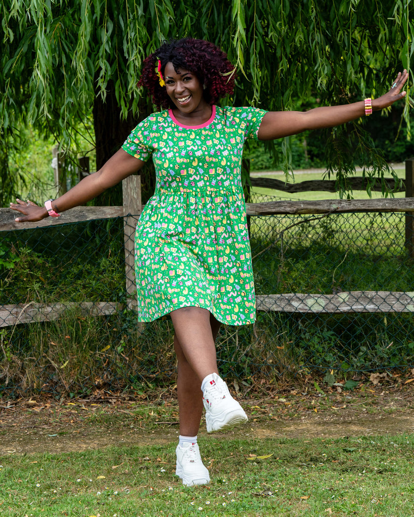 Ayesha (she/her), who is 5'10" and a UK 8/10, wears a Rebel Romance x Dipstick Store Bugs Stretch Smock Dress in size 2XS. The green smock dress features a playful multicoloured bug print with a pink neckline. Photographed outdoors in front of a rustic wooden fence and lush greenery, Ayesha poses joyfully, showcasing this ethical, alternative fashion piece for fun-loving millennials.
