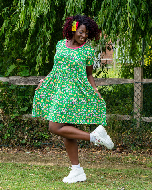 Ayesha (she/her), 5'10" and a UK size 8/10, wears the Rebel Romance x Dipstick Store Bugs Stretch Smock Dress in size 2XS. The short-sleeved, knee-length dress features a fun bug print on green stretch cotton, finished with a contrasting pink neckline. Photographed mid-pose under a weeping willow, this image captures the carefree spirit of Rebel Romance’s slow fashion for millennials.