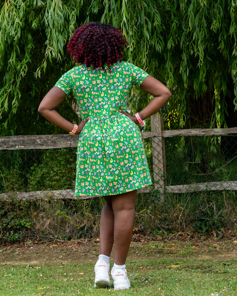 Back view of Ayesha (she/her), 5'10" and a UK 8/10, modelling the Rebel Romance x Dipstick Store Bugs Stretch Smock Dress in size 2XS. The vibrant green dress with an all-over bug print flares out from the waist, styled for movement and comfort. Shot outdoors with willow trees and wooden fencing, this photo highlights the playful and inclusive design by UK brand Rebel Romance.