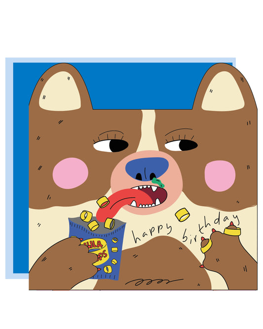 Square greeting card featuring a brown and cream dog with pink cheeks, playfully eating Hula Hoops crisps. Some Hula Hoops are on its claws, and “happy birthday” is written across the illustration. Comes with a Kraft envelope.