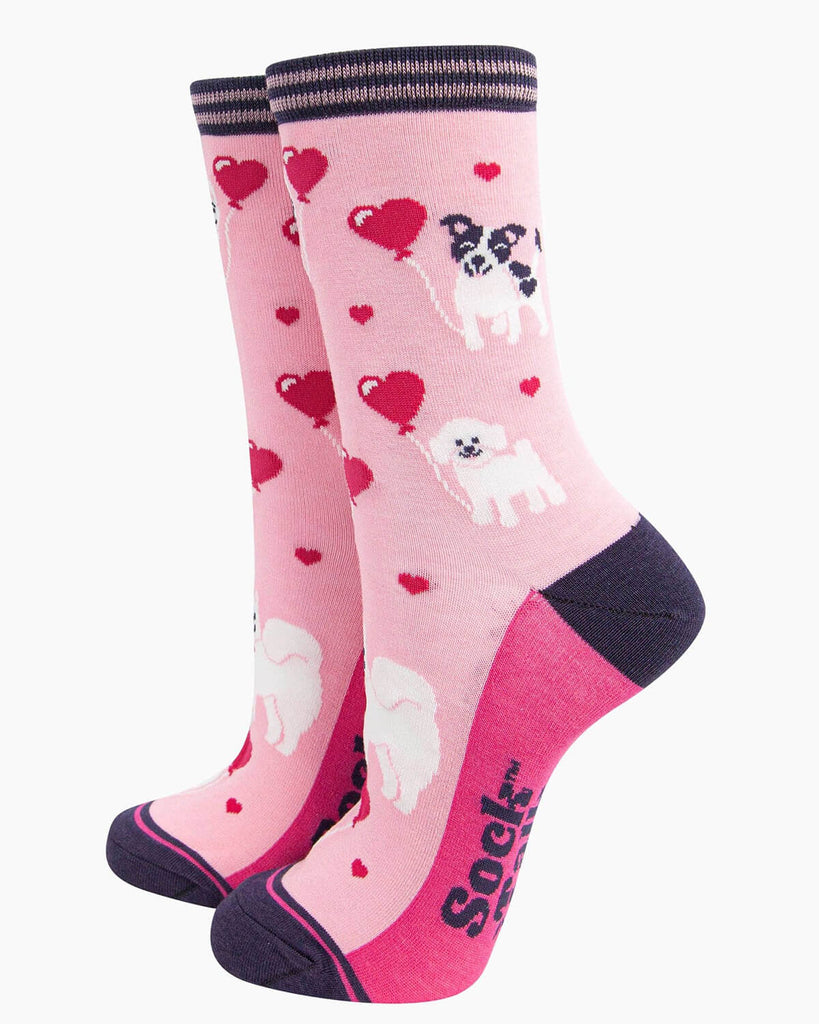 (Socks Standing Up): A pair of pink bamboo socks featuring playful dogs holding heart balloons, displayed upright. The design includes fluffy white and black-and-white pups, floating hearts, and a contrasting dark purple heel and toe.