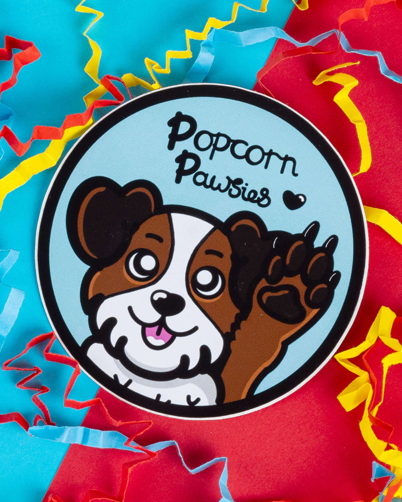 Popcorn Pawsies dog sticker by Innabox, sold by Rebel Romance. Features a cute cartoon dog with a happy face and raised paw on a light blue circular background. Surrounded by colourful confetti-style paper on red and blue backdrop. Ideal for dog lovers and fans of quirky, illustrated vinyl stickers from small UK businesses.
