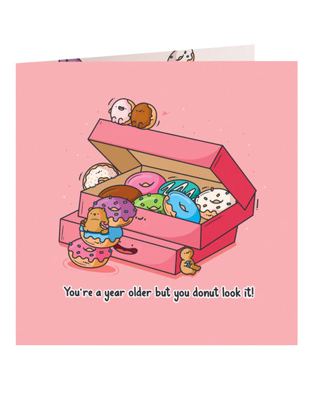 Front cover of the Toastedink birthday card featuring a pink box of colourful cartoon donuts with smiling faces and a bear climbing the donuts, with the caption "You're a year older but you donut look it!" against a pastel pink background.