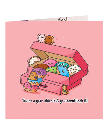 Front cover of the Toastedink birthday card featuring a pink box of colourful cartoon donuts with smiling faces and a bear climbing the donuts, with the caption "You're a year older but you donut look it!" against a pastel pink background.