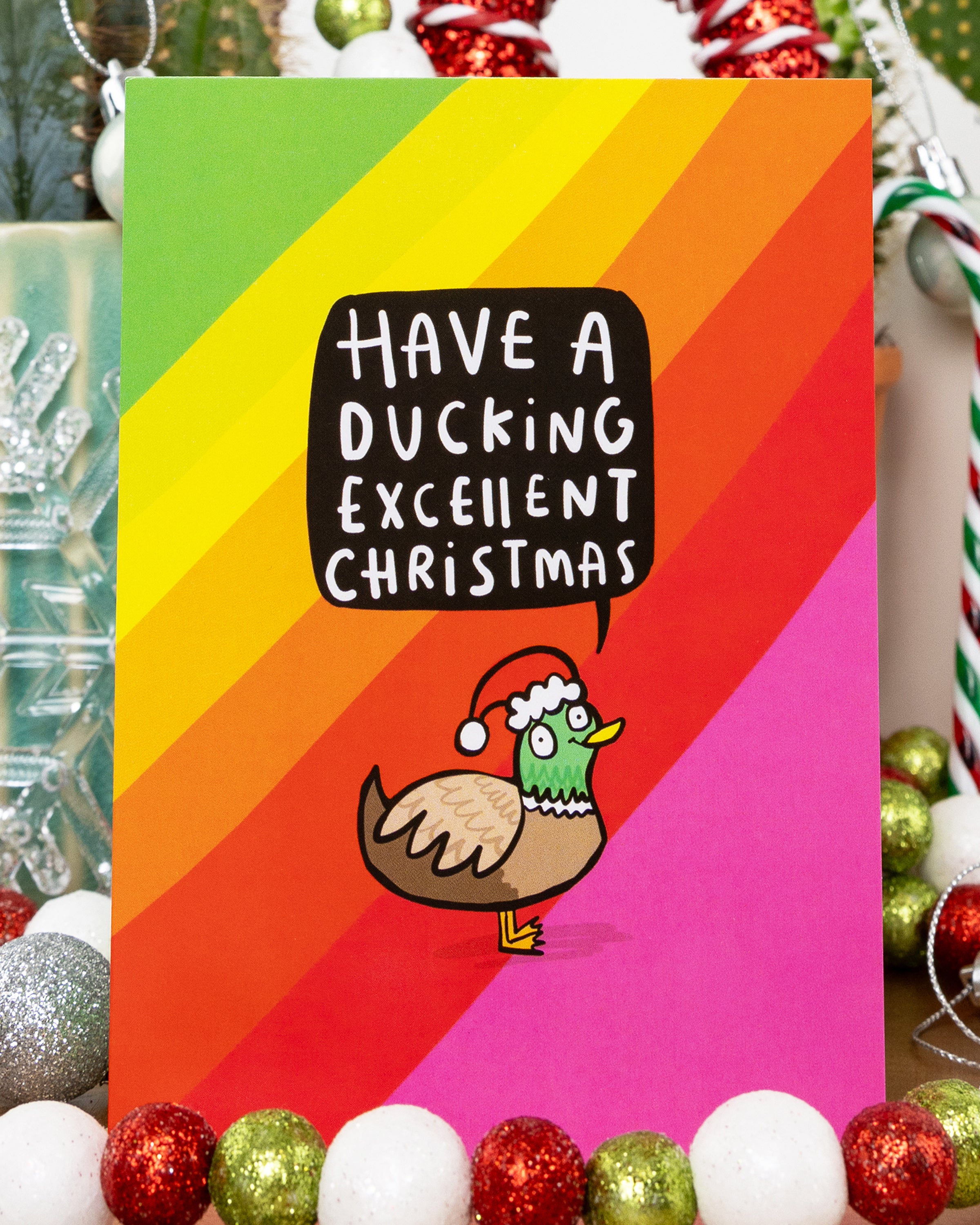 Funny Christmas card by Rebel Romance featuring a cartoon duck in a Santa hat with a speech bubble saying “Have a Ducking Excellent Christmas” on a rainbow background. Designed by Katie Abey. Surrounded by festive decor including baubles, candy canes and tinsel. A quirky and colourful Christmas greeting card perfect for millennials with a cheeky sense of humour.